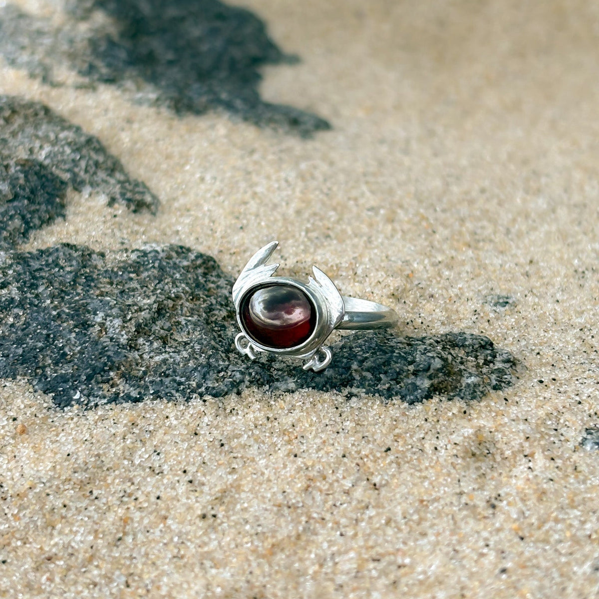 A silver ring with a round dark red cabochon gemstone, resting on a sandy beach with dark wet sand in the background.