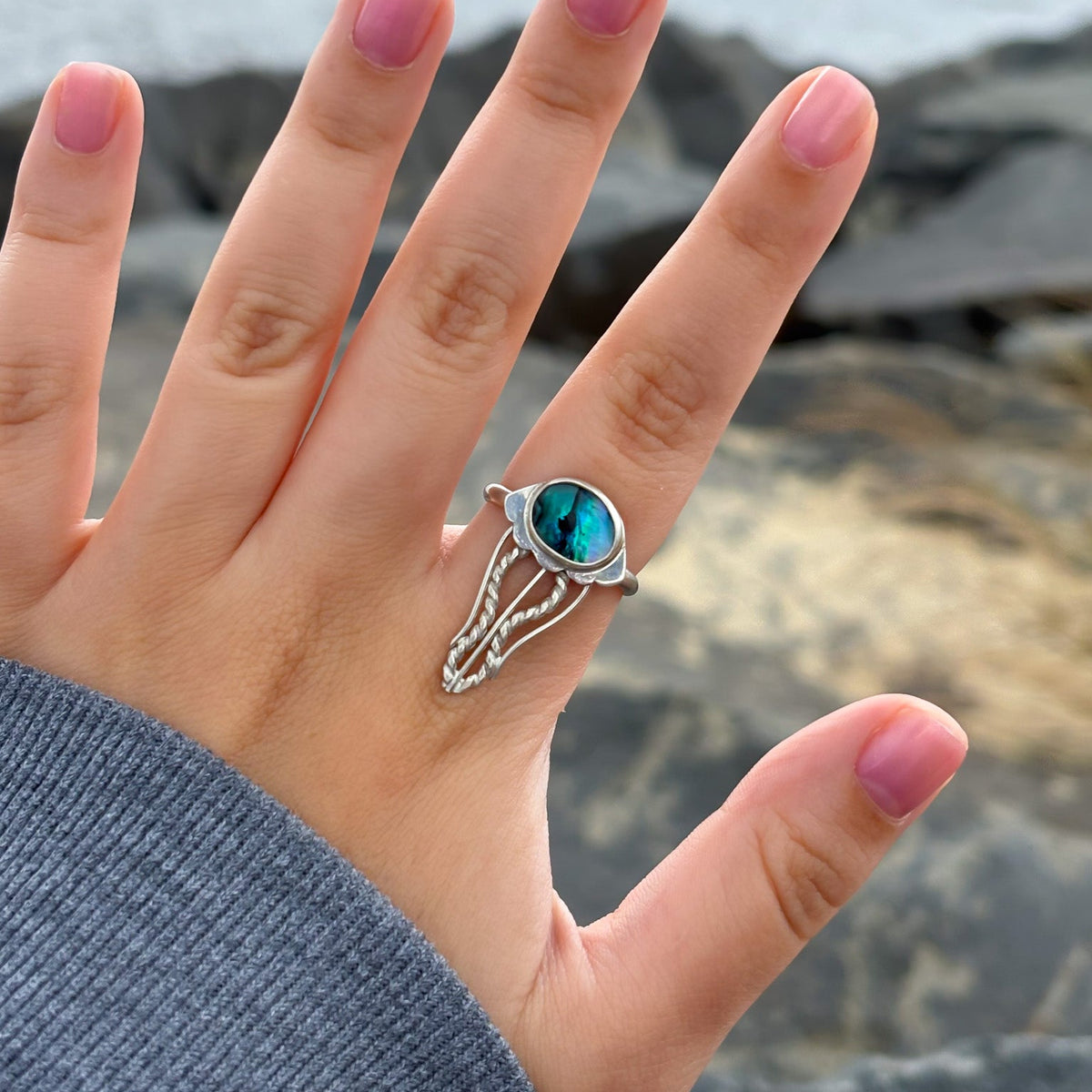 A silver ring with a round turquoise-blue stone and delicate pavé accents, shown worn on a hand with pink nail polish against a blurred rocky shoreline background.