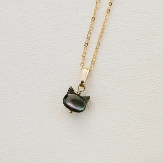 A gold chain necklace with a small cat-shaped pendant made of dark, iridescent mother-of-pearl, displayed on a textured white surface.