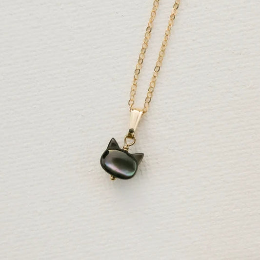 A gold chain necklace with a small cat-shaped pendant made of dark, iridescent mother-of-pearl, displayed on a textured white surface.